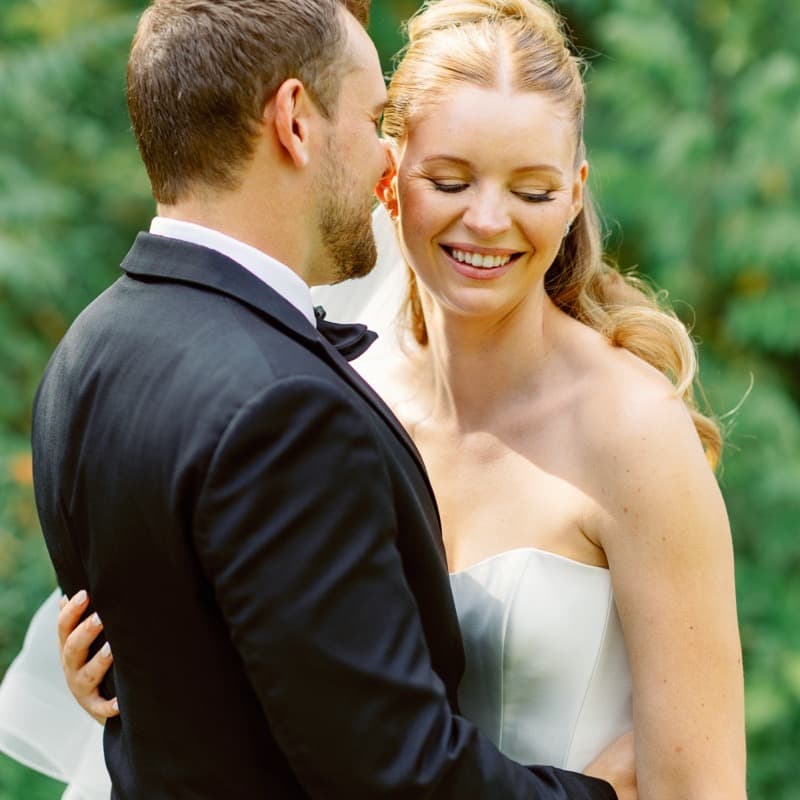 Groom whispering to smiling bride in garden light