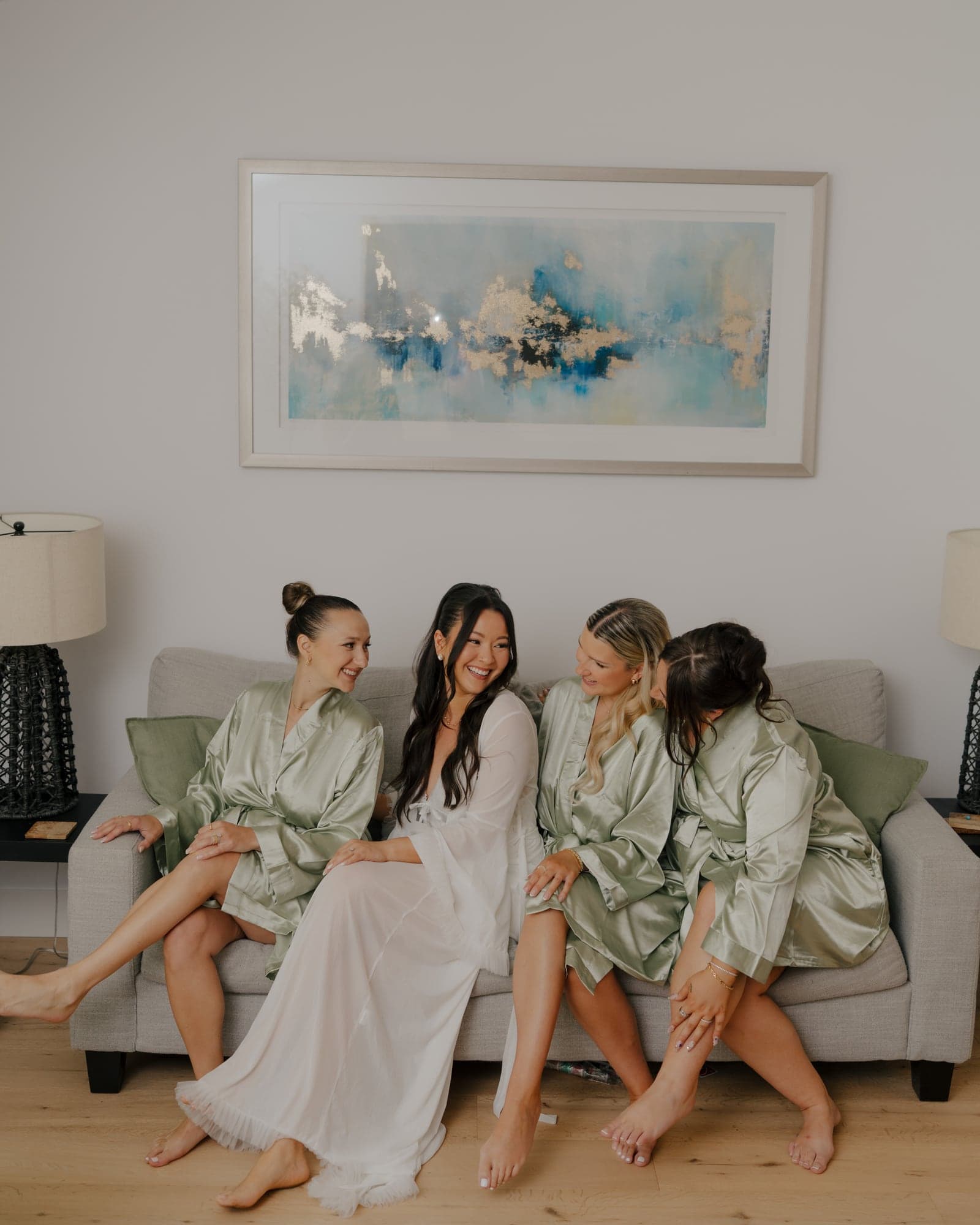Bride laughing with her bridesmaids in matching sage robes while getting ready